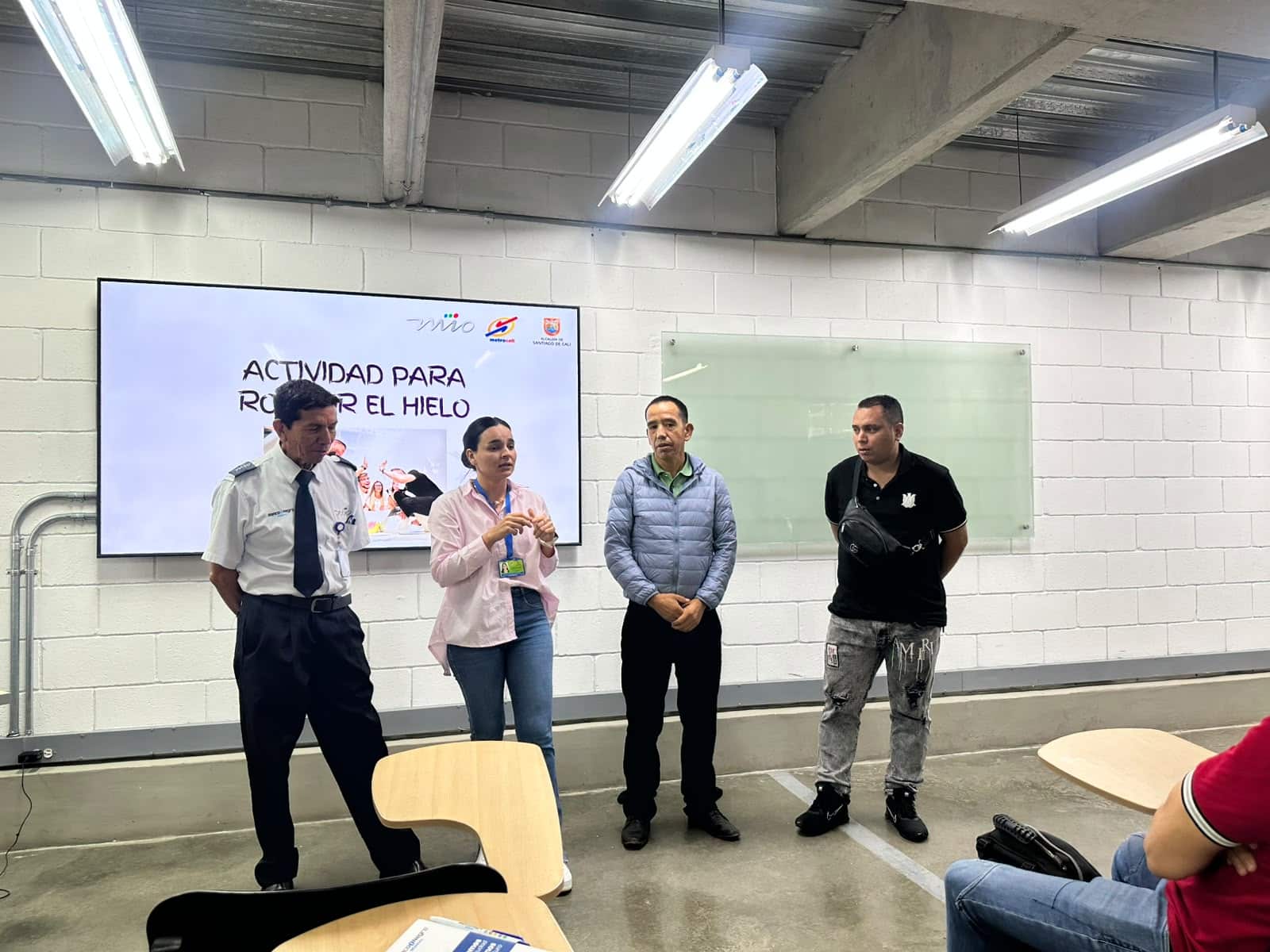 Fotografía de una capacitación en una sala. Cuatro personas están de pie al frente, incluyendo a un funcionario con uniforme, mientras una mujer explica un tema. De fondo, una pantalla proyecta el título "ACTIVIDAD PARA ROMPER EL HIELO" junto a los logos del MIO, Metro Cali y la Alcaldía.