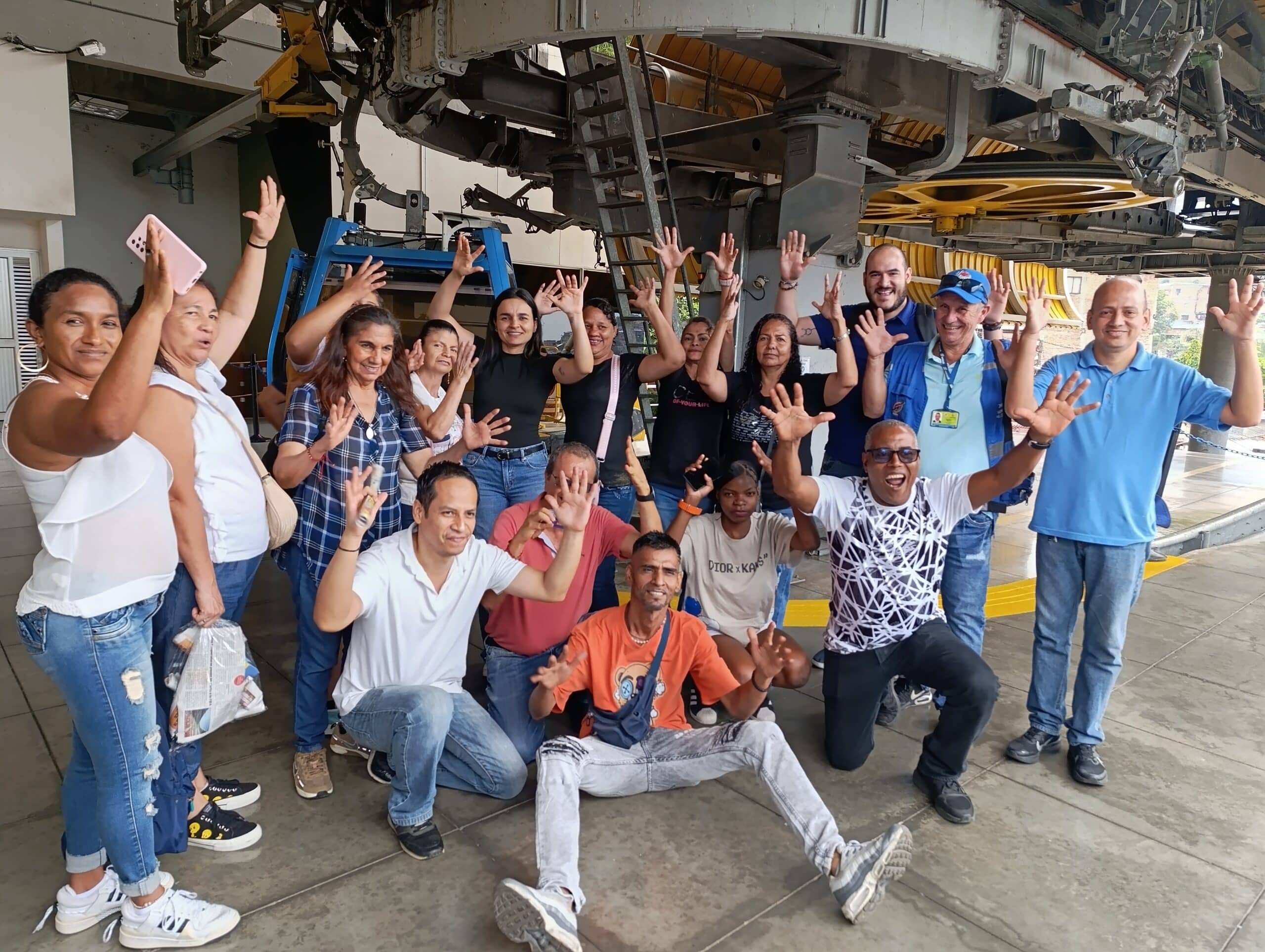 Un grupo numeroso de ciudadanos sonriendo y saludando a la cámara con las manos en alto. Se encuentran en las instalaciones de una estación, con la maquinaria y las cabinas azules del MIO Cable visibles en la parte trasera.