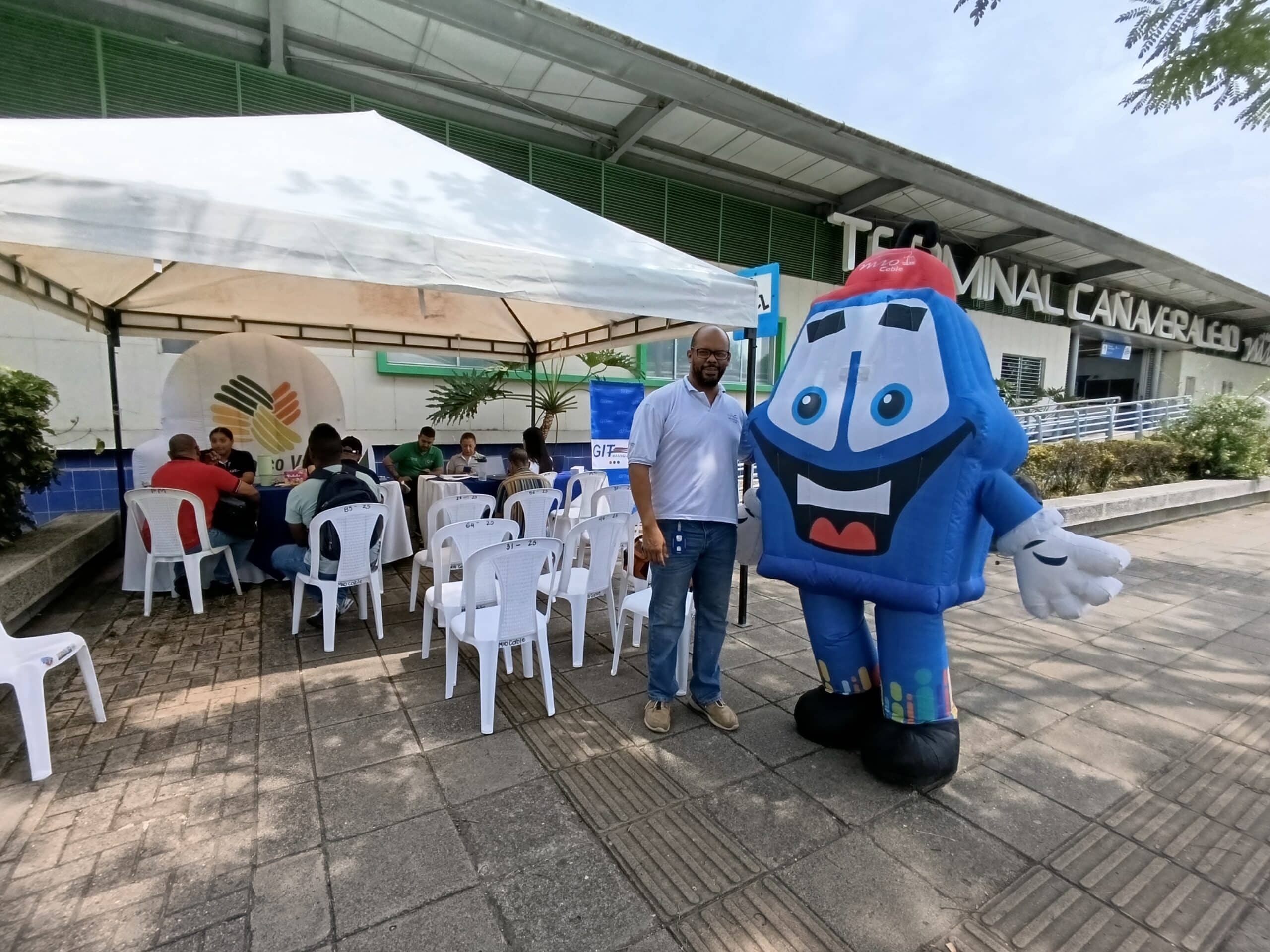 Un hombre posa sonriente junto a una gran mascota inflable del MIO de color azul y rojo. Están ubicados en los exteriores de la Terminal Cañaveralejo. Atrás se observa una carpa blanca con personas participando en una jornada de atención o evento.