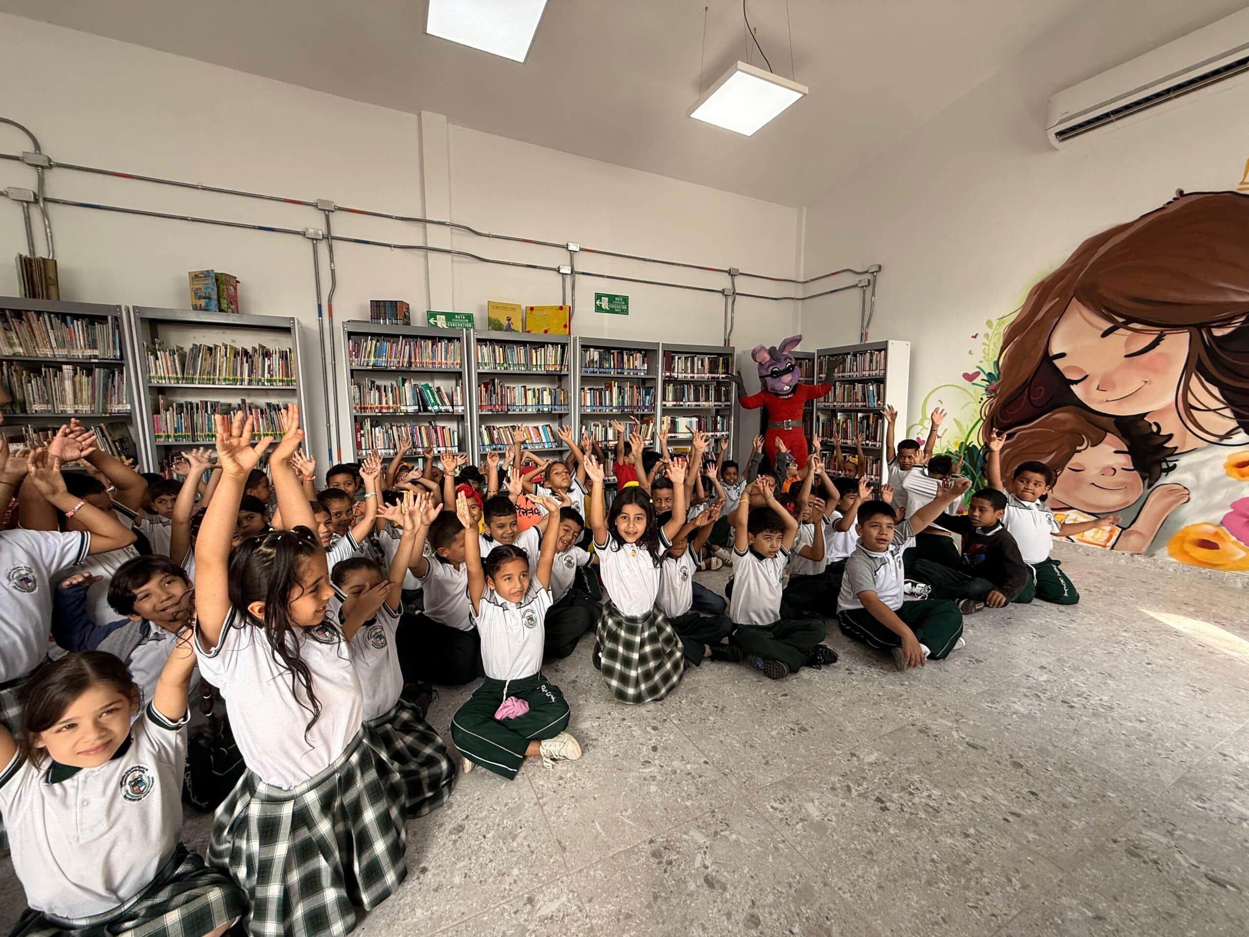 Un grupo de niños y niñas sentados en el suelo de una biblioteca escolar, levantando las manos con entusiasmo. Al fondo se observa la mascota del "Doctor Muelitas" y estanterías con libros. En la pared derecha destaca un gran mural pintado.