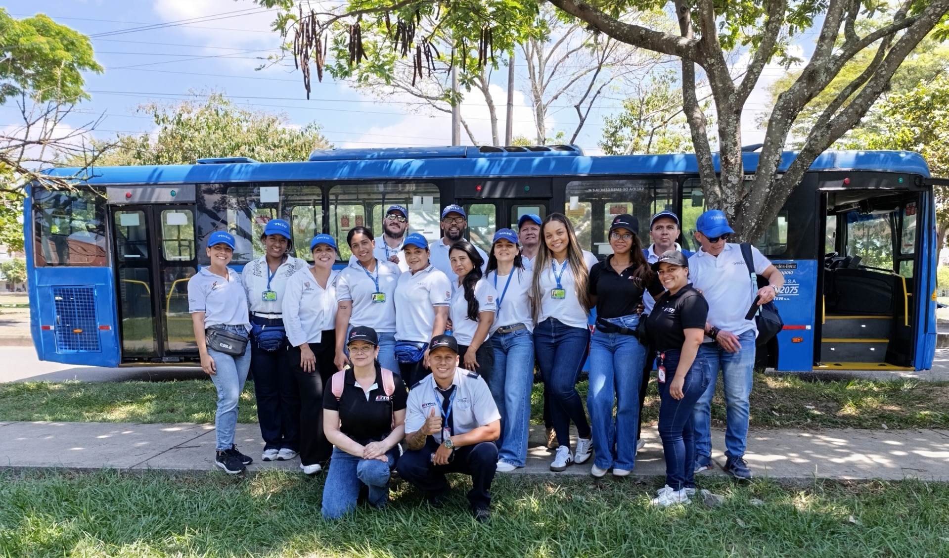Fotografía grupal de un equipo de trabajo posando frente a un bus azul del MIO. El grupo está compuesto por hombres y mujeres, la mayoría vistiendo gorras azules y camisetas con logos institucionales, mostrando un ambiente de compañerismo.