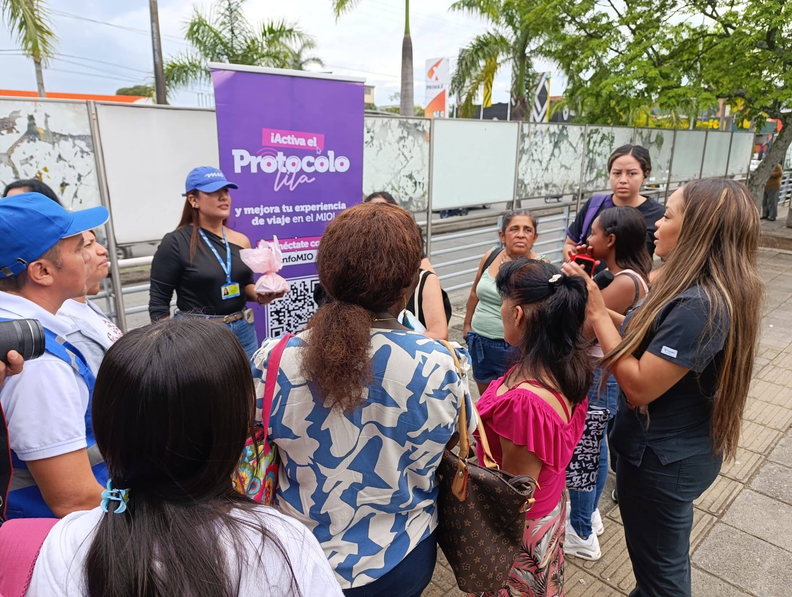 Actividad de socialización en espacio público. Un grupo de mujeres y personal de atención del sistema interactúan frente a un pendón morado de la campaña que dice "¡Activa el Protocolo Lila y mejora tu experiencia de viaje en el MIO!".