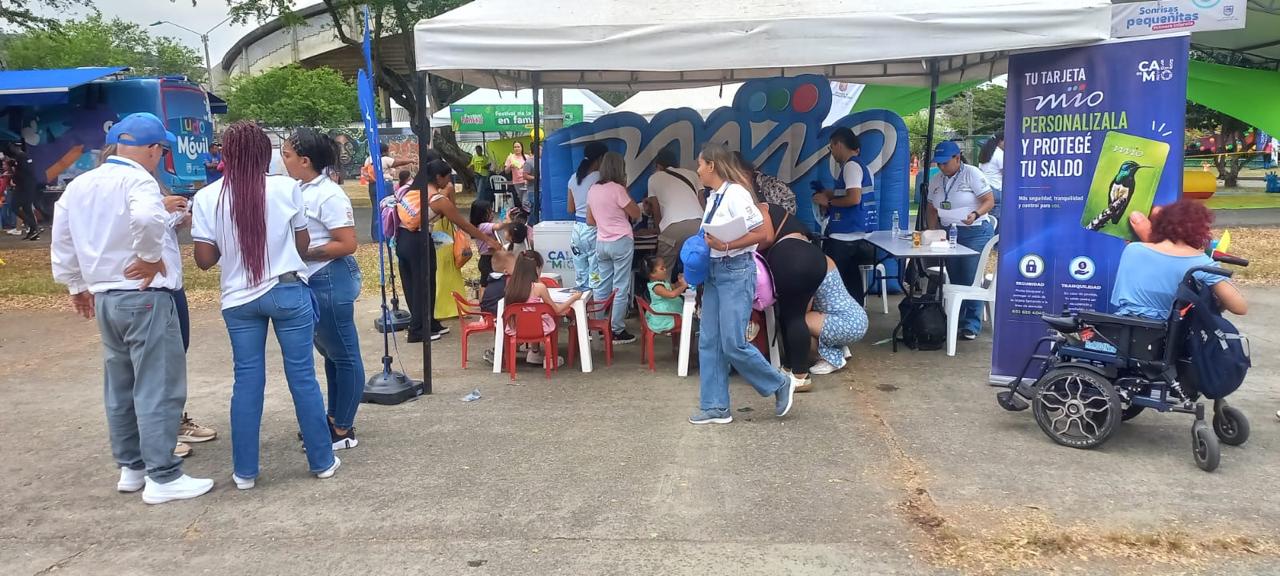 Estand de atención de Metro Cali en el Festival de la Familia. Personal con chalecos azules asiste a ciudadanos, incluyendo niños y una persona en silla de ruedas, frente a un gran inflable con el logo del MIO y un pendón promocional sobre la personalización de la tarjeta.