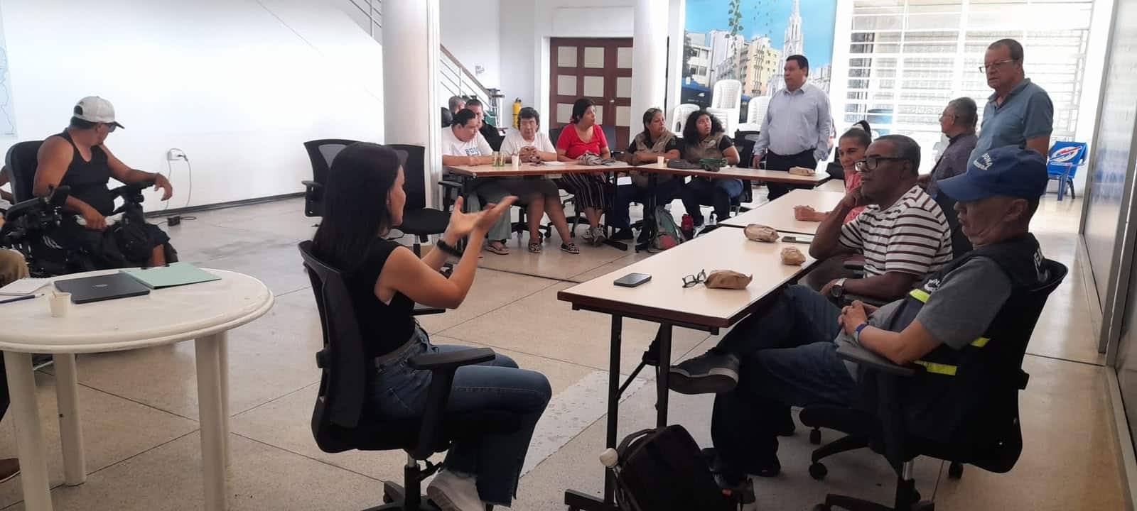 Reunión de la mesa de accesibilidad en un espacio interior cerrado. Una funcionaria de Metro Cali gesticula mientras habla frente a un grupo diverso de representantes de la comunidad con discapacidad, quienes escuchan atentamente sentados alrededor de mesas dispuestas en forma de U.