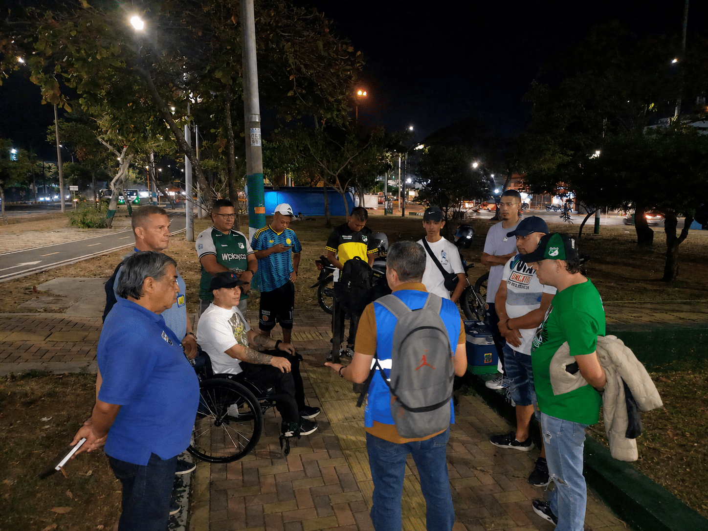 Reunión nocturna al aire libre en el Parque Estación Los Guaduales. Un funcionario con chaleco azul y mochila dialoga en círculo con un grupo de jóvenes barristas, algunos vistiendo camisetas del Deportivo Cali y uno de ellos en silla de ruedas, durante la mesa de trabajo sobre el cuidado de la infraestructura.