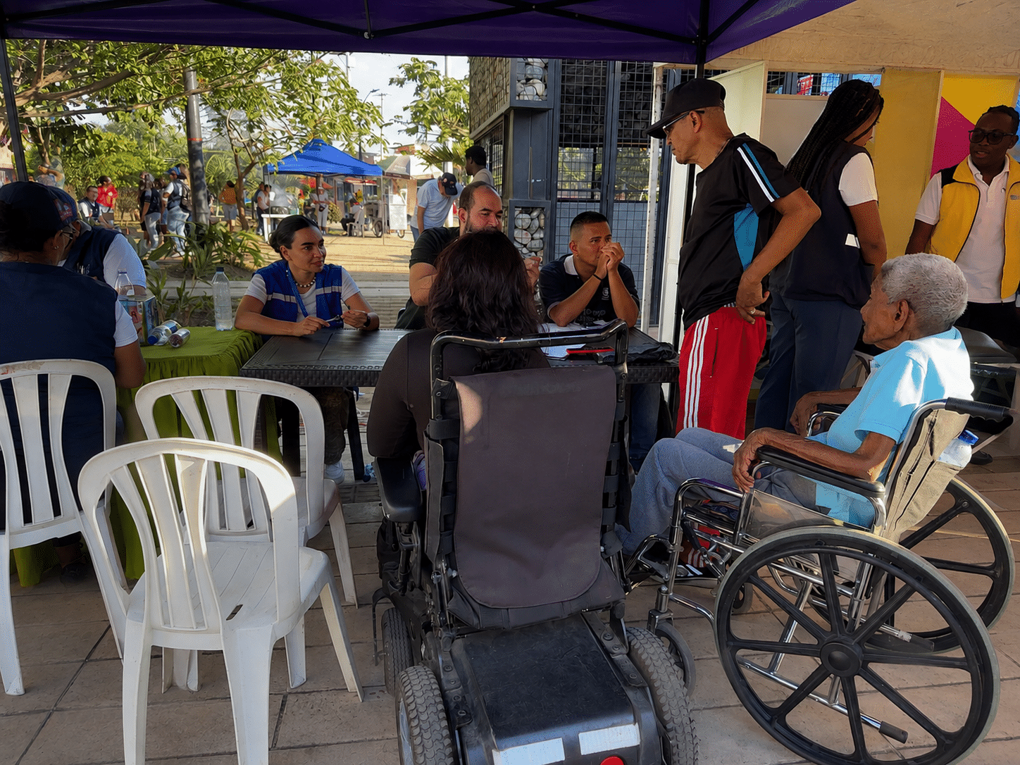 Punto de atención al aire libre bajo una carpa morada. Funcionarios del distrito y de Metro Cali asisten en unas mesas a varias personas, incluyendo a una adulta mayor en silla de ruedas, durante la jornada de verificación y entrega de tarjetas de acceso preferencial Puerta Glass.