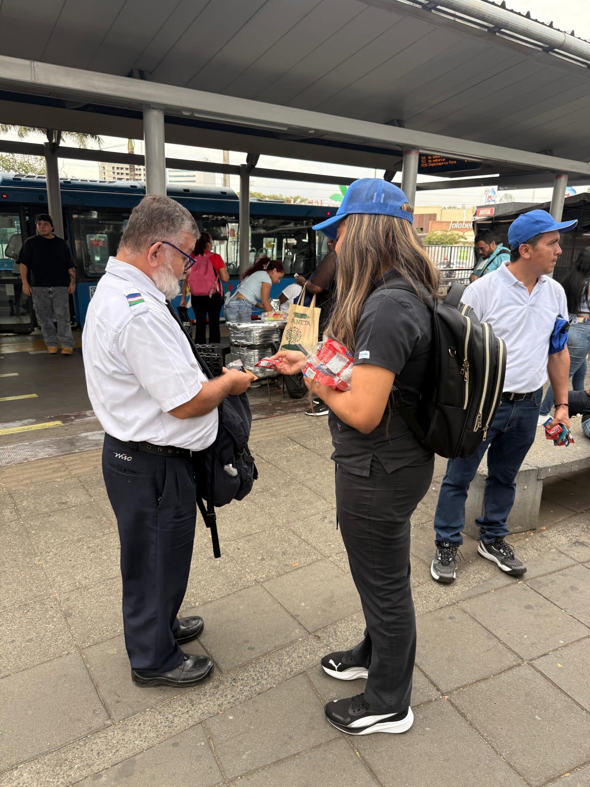 Una colaboradora de Metro Cali entrega un obsequio a un operador de bus del MIO en la plataforma de una estación. El conductor viste su uniforme blanco con el logo del sistema, mientras que al fondo se observa un bus azul estacionado, en el marco de la celebración del Día del Conductor.