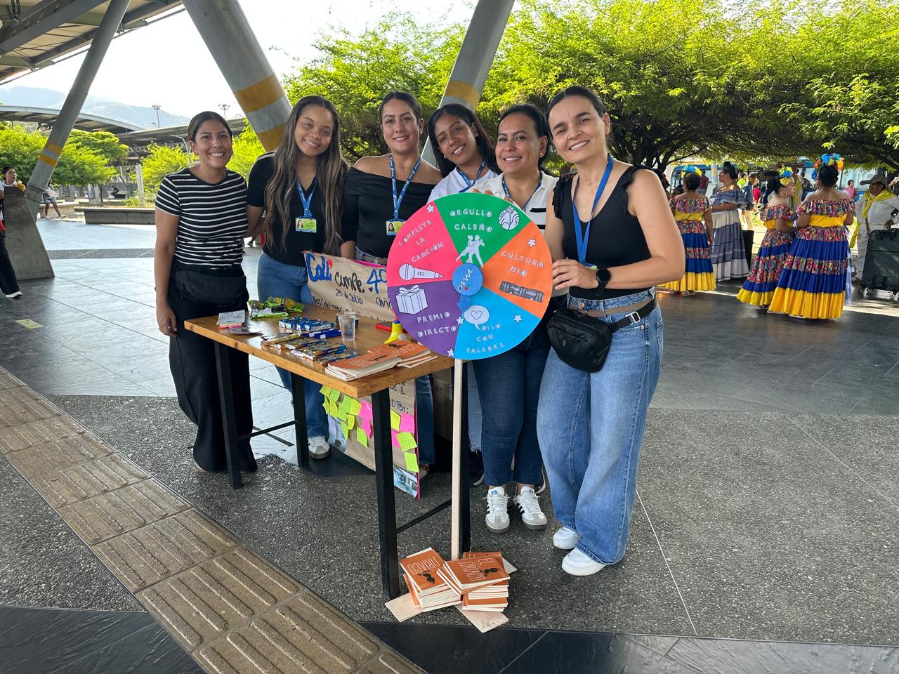 Grupo de seis mujeres sonriendo detrás de una mesa de atención que exhibe dulces y una colorida ruleta interactiva con temáticas de ciudad y cultura MIO. Al fondo de la terminal, se observan personas vistiendo faldas y trajes típicos por la celebración de los 490 años de Cali.