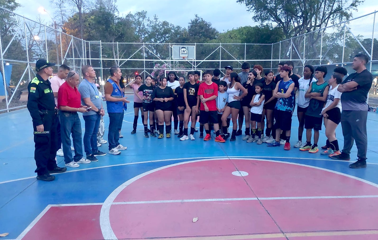 Jornada comunitaria en una cancha deportiva al aire libre al atardecer. Un funcionario de Metro Cali con chaleco azul y un policía de Colombia están a la izquierda de la imagen, hablando con un gran grupo de adolescentes y niños vestidos con ropa deportiva que están de pie en semicírculo frente a ellos. El suelo de la cancha está pintado de azul y rojo, y está rodeada por una alta valla de malla de alambre.