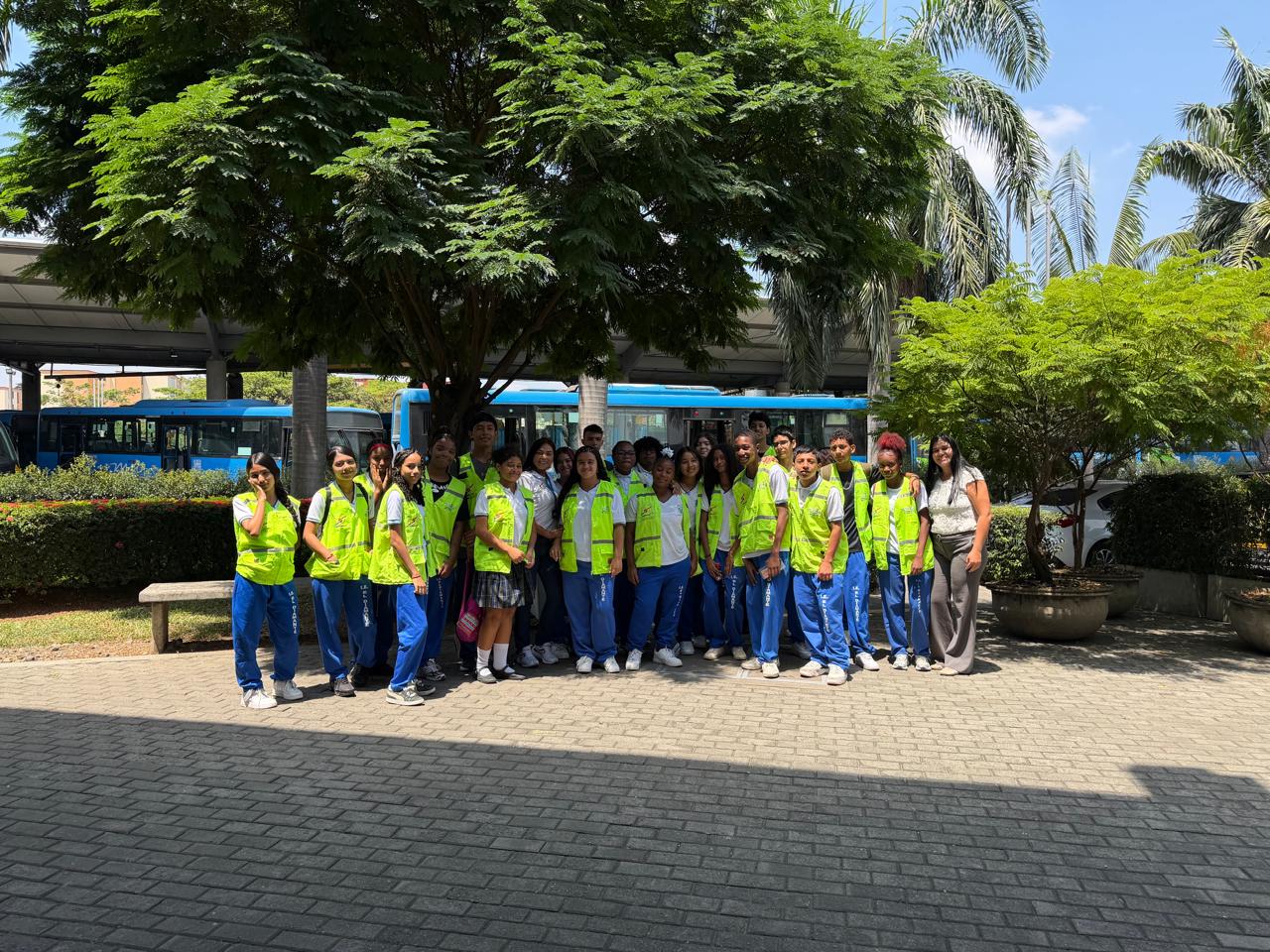 Grupo numeroso de estudiantes de la Institución Educativa El Diamante posando sonrientes bajo un árbol. Los jóvenes visten su uniforme escolar junto con chalecos reflectivos verdes del programa Escuela MIO. De fondo se observa la infraestructura de la terminal y varios buses azules del sistema.